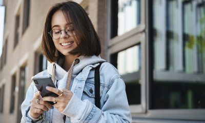 young queer girl college student standing street order taxi using application smartphone smiling as texting friend meeting with team outside campus scrolling news feed social media 1